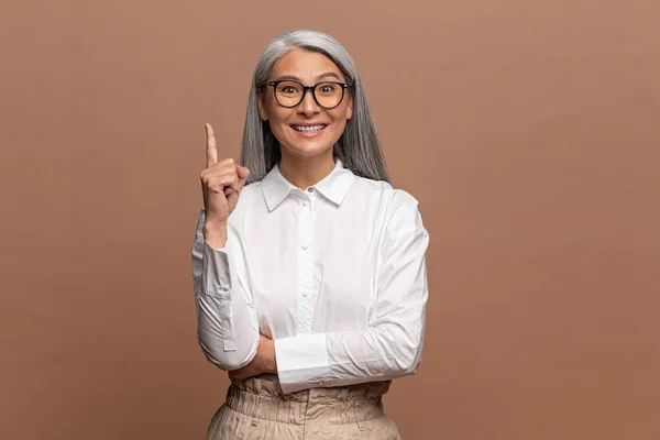 Mujer madura hermosa asiática con gafas y ropa formal se le ocurrió la idea y levantó el dedo. Estudio aislado filmado en beige — Foto de Stock