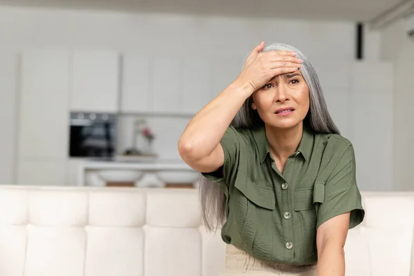 Занепокоєна сиве волосся жінка тримає лоб з рукою, сидячи на дивані вдома — стокове фото