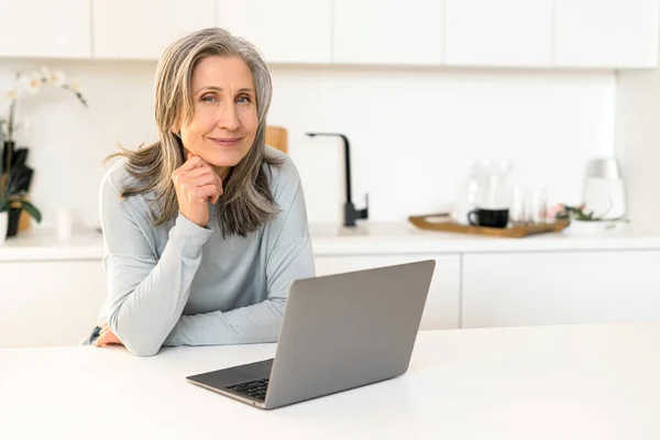 Gai attrayant mature femme regarde assis dans la cuisine avec ordinateur portable — Photo