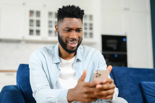 Fokuserat skägg multiracial man sitter på soffan hemma och håller smartphone. Killen läser nyheter på nätet, tillbringar fritid i sociala nätverk, mörkhårig manlig sms och chatta — Stockfoto