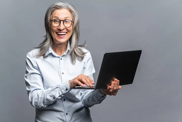 Сучасна літня жінка в офіційному вбранні з використанням ноутбука ізольована на сірому. Портрет зрілої жінки-офісу працівника, що використовує онлайн-технології для ведення бізнесу — стокове фото