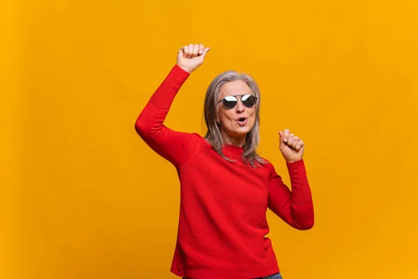 Concepto de humor navideño. Mujer alegre y alegre de pelo gris levantando brazos y bailando. Señora madura con gafas de sol celebrando algo aislado en amarillo — Foto de Stock