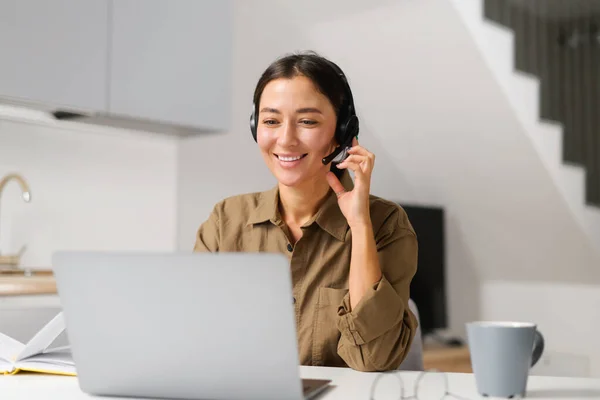Joven mujer alegre usando auriculares para hablar en línea sentado en el escritorio en la oficina en casa. Agradable operador femenino mirando a la pantalla — Foto de Stock