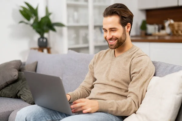 Веселый кавказский мужчина в повседневной одежде сидит на диване и пользуется ноутбуком в современном домашнем офисе. Печатание на клавиатуре — стоковое фото