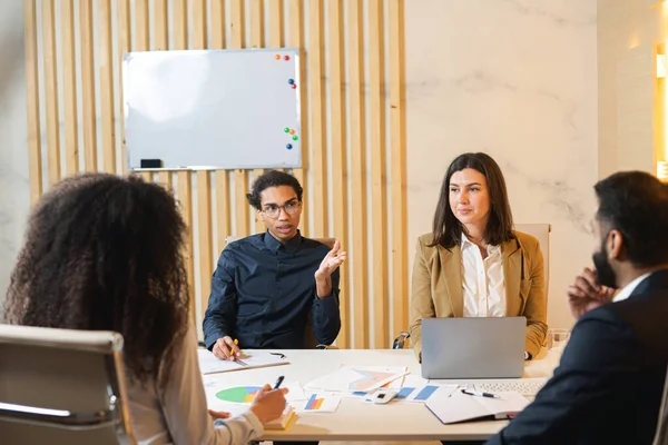 Friendly cheerful atmosphere at the business meeting of a multiracial young team. Young guy telling something to his colleagues on the meeting at the office — Stock Photo, Image