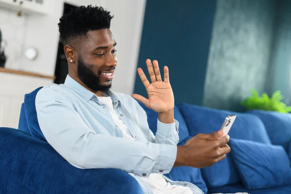 Spännande ung multiracial man sitter på soffan och använder smartphone för videoanslutning, viftande hand, hälsning vän eller familj på avstånd — Stockfoto