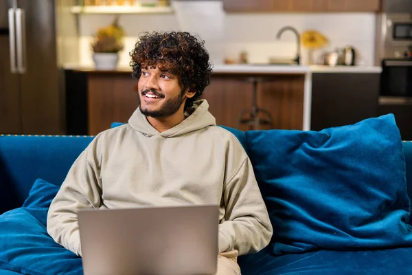 Optimistický etnický muž v neformálním oblečení používá notebook, když sedí na gauči. Mladý indický student sledující webináře, vzdělávací kurzy, učení na dálku — Stock fotografie