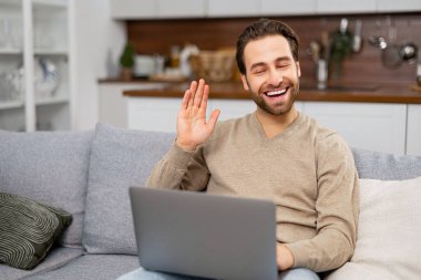 Neşeli kaygısız adam video bağlantısı için dizüstü bilgisayar kullanıyor, webcam 'e bakıyor ve el sallıyor. Adam kanepede oturuyor ve video görüşmesi yapıyor.