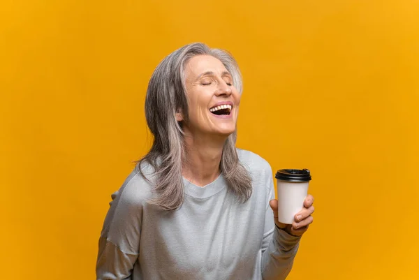 Mujer mayor sonriente en ropa casual sosteniendo taza de papel de café aislado sobre fondo amarillo. Madura de pelo gris dama de pie y riendo, mientras disfruta del café para llevar — Foto de Stock