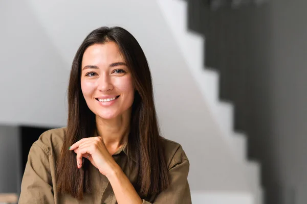 Porträt einer charmanten, eleganten asiatischen Frau mit der Hand unterm Kinn, die in die Kamera lächelt, im modernen Haus sitzend, in häuslicher Kleidung — Stockfoto