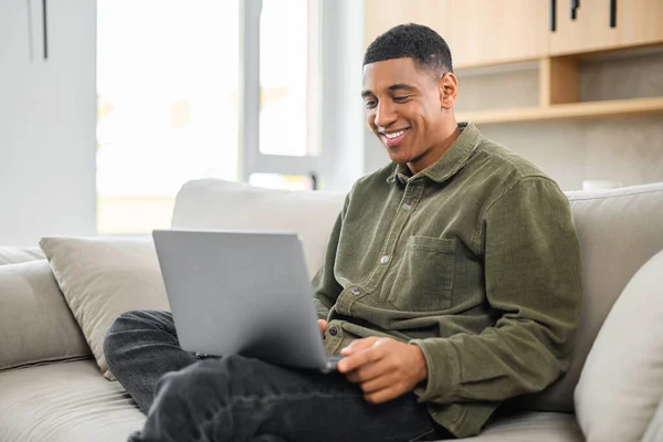 낙관적 인 사람은 소파에 앉아 있는 동안 노트북을 사용하여 평상 시대의 옷을 입는다. 젊은 남자 학생 이 웹툰을 보고, 교육 과정을 보고, 멀리서 배우고, — 스톡 사진