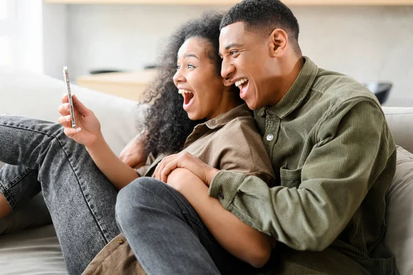 Задоволена і радісна багаторасова молода пара сидить на дивані вдома і тримає смартфон. Чоловік і жінка святкують хороші новини або перемагають — стокове фото