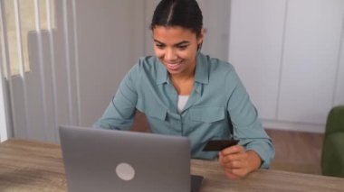 Gülümseyen, çok kültürlü bir kadın kredi kartı tutuyor ve dizüstü bilgisayar kullanıyor.