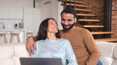 Hintli çift rahat oturma odasında kanepede oturuyor ve sanal bağlantı için laptop kullanıyorlar.