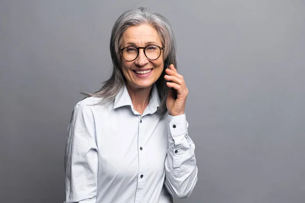 Mujer de negocios sonriente y positiva hablando con el teléfono inteligente sobre la pared gris. Vendedora de ancianos tiene conversación telefónica. Madura mujer hablando en el teléfono móvil — Foto de Stock