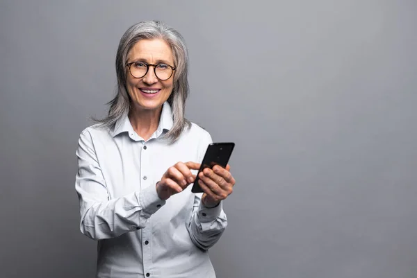 Šťastná obchodní žena ve formální košili dotýká obrazovky smartphone a zároveň se cítí skvěle s novými funkcemi telefonu. Vnitřní studio záběr izolované na šedé — Stock fotografie