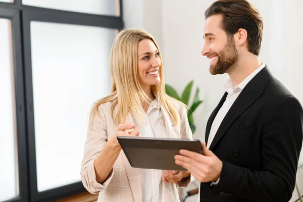 Jeune femme en chemise blanche est assise à la table au bureau et regardant son collègue masculin tout en tenant la tablette avec le sourire — Photo