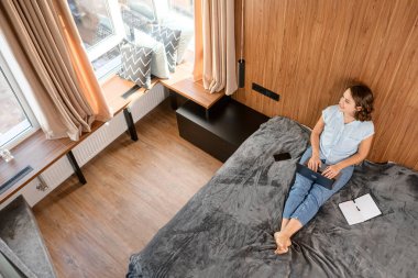 Yatakta oturmuş, dizüstü bilgisayarında e-posta yazan, rahat giysiler içindeki genç iş kadını ya da kadın serbest çalışanın yüksek açılı görüntüsü.