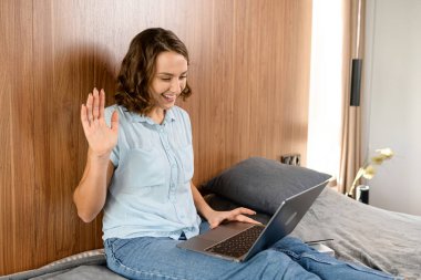 Beyaz kadın, yatakta otururken akıllı telefonu video bağlantısı olarak kullanıyor. Gülümseyen kız el sallıyor merhaba, selam veren arkadaş ya da aile