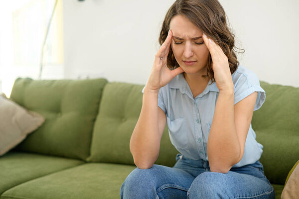 Upset young woman has strong headache, sitting on sofa at home and massaging temples, feels tired and exhausted. Sad lady worrying, feels anxiety about problems