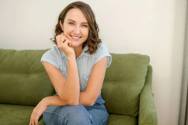Healthy woman with brunette hair with glowing eyes, sitting on the pure comfortable sofa at modern apartment, dressed in casual clothes — Stock Photo, Image
