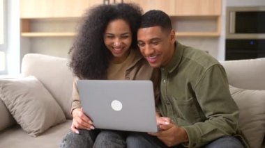 Çok ırklı bir çift, rahat oturma odasındaki kanepede otururken video araması için dizüstü bilgisayar kullanıyor.