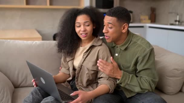 Молода багаторасова пара використовує ноутбук, сидячи на дивані вдома — стокове відео