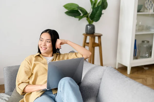 Aziatische jonge vrouw besteedt thuis vrije tijd met laptop. Rustig meisje zit op een gezellige bank en kijkt weg voor het laptopscherm. Surfen op het web, tv-series kijken — Stockfoto