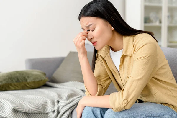 Üzgün Asyalı kadın kendini kötü hissediyor, evdeki kanepede oturuyor, başını tutuyor ve gözlerini kapatıyor. Üzgün Asyalı kadın depresyonda.