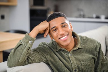 Evdeki modern oturma odasında dinlenen yakışıklı genç adamın portresi, kanepede oturmuş, kameraya bakıyor, rahatlamış hissediyor.