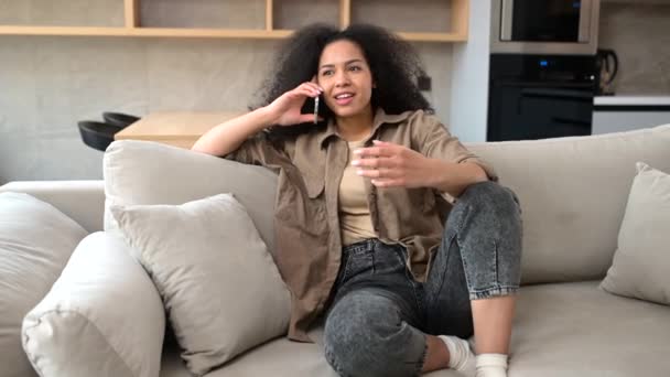 Gioioso multirazziale giovane donna riccia parlando smartphone seduto sul divano — Video Stock