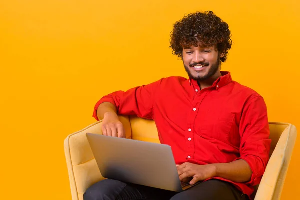 Felice uomo indiano guardando lo schermo del dispositivo, digitando contenuti informativi, utilizzando il computer portatile per lavorare, seduto sulla poltrona isolato su sfondo arancione — Foto Stock
