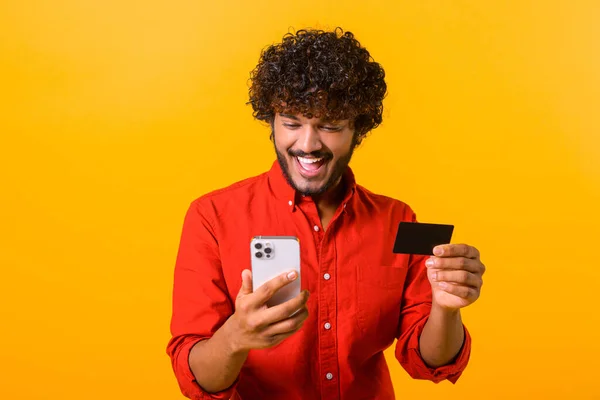 Alegre chico de Oriente Medio está utilizando el teléfono inteligente y tarjeta de crédito para ir de compras en línea. El hombre feliz está pidiendo comida en línea, el hombre paga por la tan esperada compra en el teléfono móvil — Foto de Stock