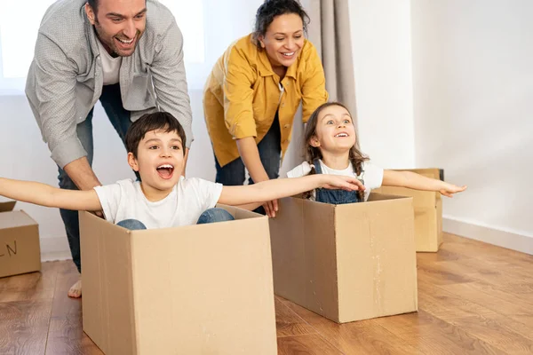 Family of four have fun in new house, parents riding kids in cardboard boxes