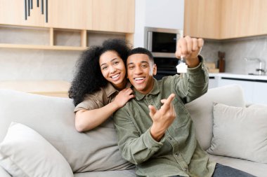 Genç evli çift neşeyle gülümsüyor ve yeni dairenin anahtarlarına bakıyor. Eşler, modern yeni evlerinde kanepede otururken kameraya sarılıp bakıyorlar.