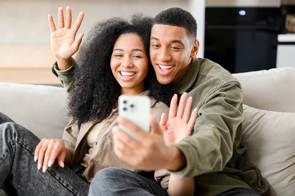 Вид спереду щаслива весела молода багаторасова пара, що має онлайн-веб-чат з через відеодзвінок на мобільний телефон, сидячи разом у затишній квартирі — стокове фото