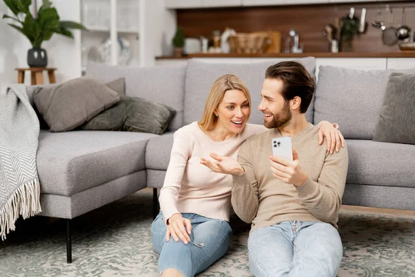 Радісна і радісна кавказька молода пара сидить біля дивана вдома і тримає смартфон. Чоловік і жінка можуть вірити в щось, святкуючи хороші новини або виграти — стокове фото