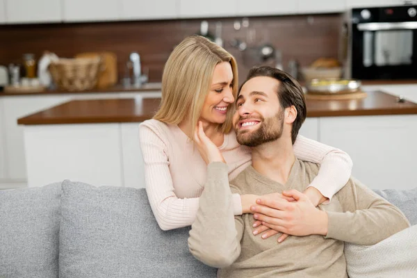 Felice bella moglie che abbraccia dalla schiena suo marito seduto sul divano, si guardano e sorridono — Foto Stock