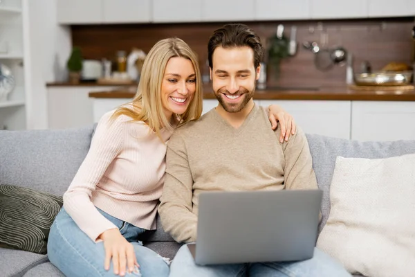 Блондинка і чоловік дивляться на екран ноутбука, сидячи на дивані і дивлячись комедійні фільми. Закохана пара проводить дозвілля онлайн — стокове фото