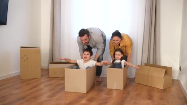 Multiracial familj på fyra ha kul i nytt hus, föräldrar ridning barn i pappkartonger — Stockvideo
