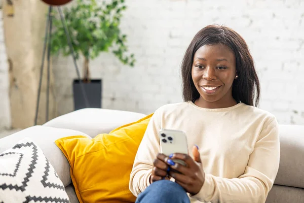 Midjan upp porträtt av den unga leende multiracial kvinna sitter vid soffan hemma och använder smartphone, fick oväntade goda nyheter — Stockfoto