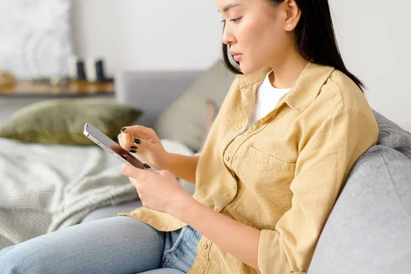 Seitenansicht der Asiatin, die auf ihr Smartphone in der Hand blickt. Weibchen telefonieren im Sitzen auf dem Sofa — Stockfoto