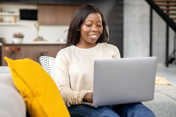Multiracial charmig ung kvinna sitter på en soffa i vardagsrummet, tittar på laptop, ler, chatta, njuta av fritid helg hemma, på internet — Stockfoto