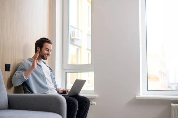 Hilarische blanke man met behulp van laptop voor externe videoverbinding, vrolijke man houdt virtuele vergadering, zitten op de bank in modern appartement, kijkt naar webcam en zwaaien — Stockfoto