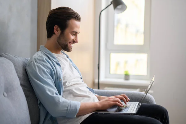 Optimistische man die op de bank zit en laptop computer gebruikt als berichtje. Gelukkige zakenman werkt op afstand in ontspannen houding — Stockfoto