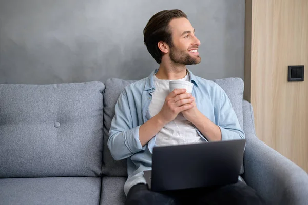 Charismatische blanke man zittend op de bank met een laptop op zijn schoot, wegkijkend en glimlachend. Freelancer man houdt van werken op afstand — Stockfoto