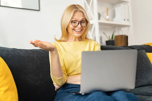 Позитивная зрелая белая белая фрилансер в очках сидит на диване с ноутбуком на коленях, учится на вебинаре онлайн или курс дома — стоковое фото
