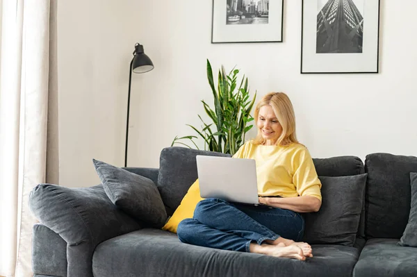 Mulher madura caucasiana passar lazer em casa com um laptop. Senhora sorrindo sentado no sofá aconchegante e olhando para a tela do laptop. Conceito de navegação na Web — Fotografia de Stock