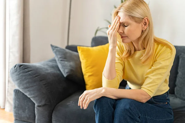 Tired mature woman sitting on the sofa at home, rubbing temple with suffering face, feels sick and weary, senior female suffer from strong headache, feels unwell, has trouble