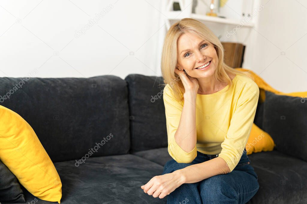 Portrait on blonde-haired woman in home interior. Charming mature caucasian lady looking at camera and smiles, resting, serene female spending weekend